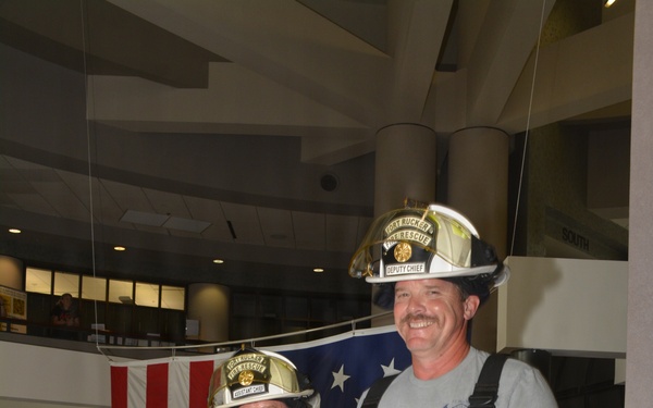 Two firefighters from Fort Rucker Fire &amp; Rescue are posing on a staircase inside a building, smiling at the camera. The firefighter on the left wears full turnout gear with an MSA SCBA unit and a helmet labeled 'Assistant Chief,' while the one on the right wears partial gear with a white helmet labeled 'Deputy Chief.' An American flag is displayed on the wall behind them.