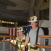 Two firefighters from Fort Rucker Fire &amp; Rescue are posing on a staircase inside a building, smiling at the camera. The firefighter on the left wears full turnout gear with an MSA SCBA unit and a helmet labeled 'Assistant Chief,' while the one on the right wears partial gear with a white helmet labeled 'Deputy Chief.' An American flag is displayed on the wall behind them.