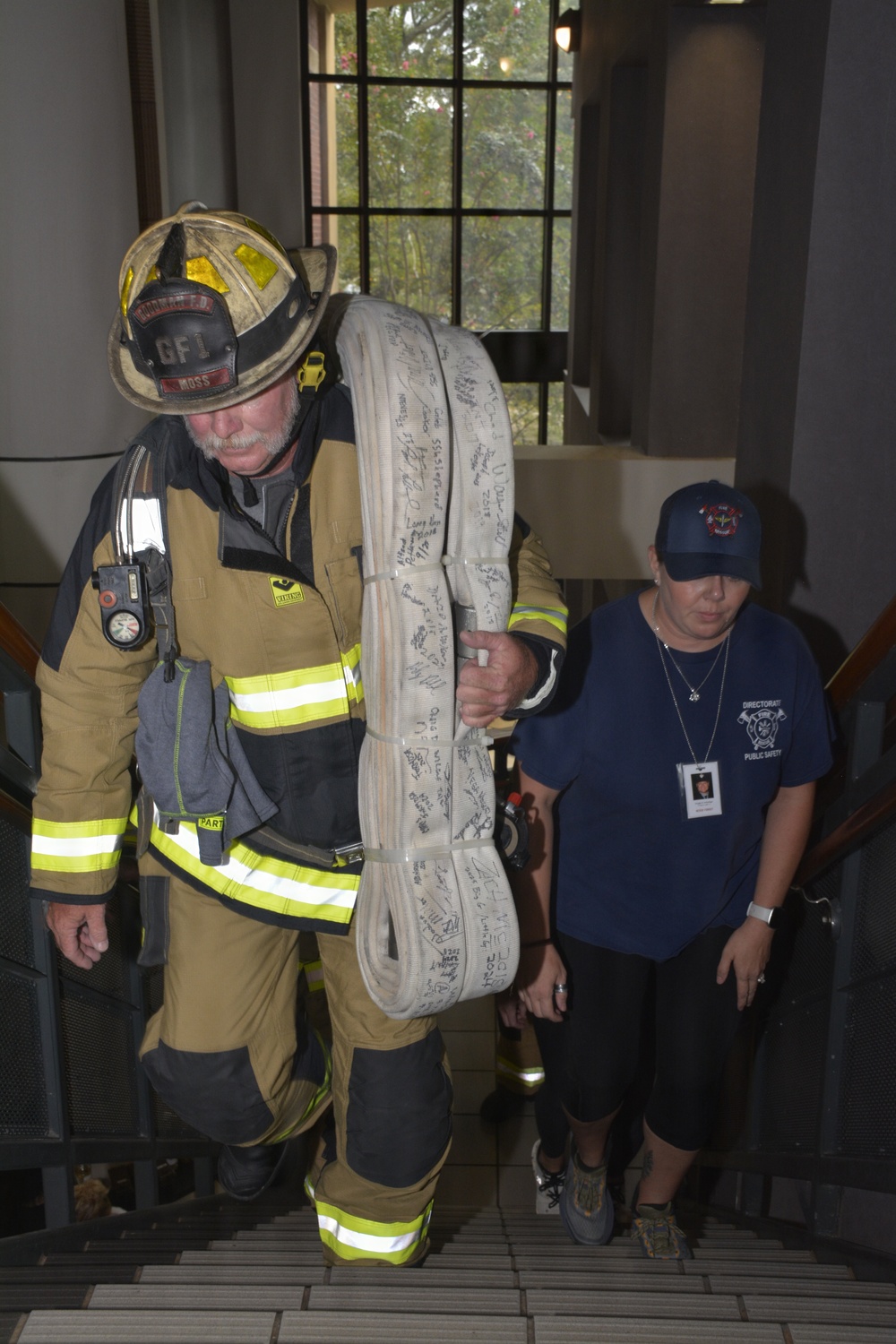 A firefighter in full turnout gear, wearing a helmet labeled 'GF1' and 'MOSS', carries a large coiled fire hose covered in signatures while climbing a staircase. Behind him, a woman in a navy blue 'Directorate of Public Safety' shirt and cap follows up the stairs wearing an ID badge. This appears to be a memorial stair climb event, likely a 9/11 tribute based on the signed hose and ceremonial nature of the activity.