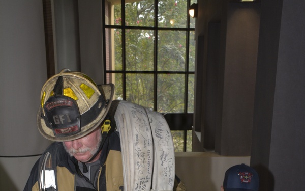 A firefighter in full turnout gear, wearing a helmet labeled 'GF1' and 'MOSS', carries a large coiled fire hose covered in signatures while climbing a staircase. Behind him, a woman in a navy blue 'Directorate of Public Safety' shirt and cap follows up the stairs wearing an ID badge. This appears to be a memorial stair climb event, likely a 9/11 tribute based on the signed hose and ceremonial nature of the activity.