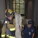 A firefighter in full turnout gear, wearing a helmet labeled 'GF1' and 'MOSS', carries a large coiled fire hose covered in signatures while climbing a staircase. Behind him, a woman in a navy blue 'Directorate of Public Safety' shirt and cap follows up the stairs wearing an ID badge. This appears to be a memorial stair climb event, likely a 9/11 tribute based on the signed hose and ceremonial nature of the activity.