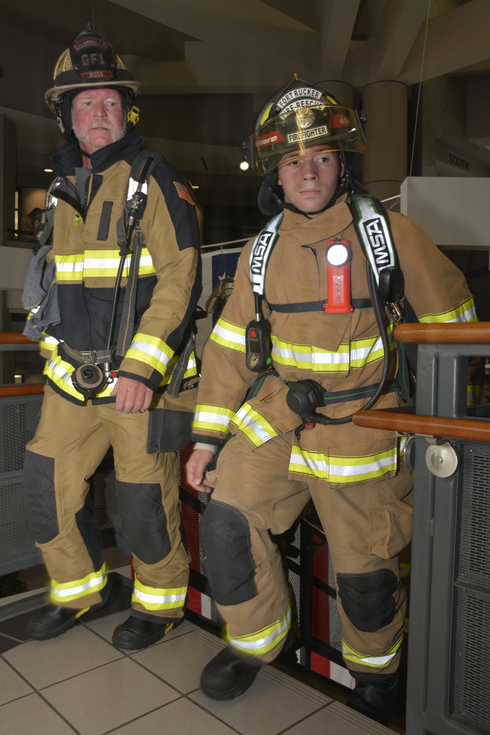 Two firefighters in full turnout gear are posed together on what appears to be a stairwell landing inside a building. The firefighter on the left wears a helmet marked 'GFI' and 'MOSS', while the one on the right wears a helmet reading 'Fort Rucker Fire-Rescue' and 'Firefighter'. Both are equipped with SCBA (Self-Contained Breathing Apparatus) gear and MSA-branded equipment.