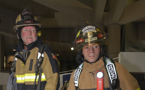 Two firefighters in full turnout gear are posed together on what appears to be a stairwell landing inside a building. The firefighter on the left wears a helmet marked 'GFI' and 'MOSS', while the one on the right wears a helmet reading 'Fort Rucker Fire-Rescue' and 'Firefighter'. Both are equipped with SCBA (Self-Contained Breathing Apparatus) gear and MSA-branded equipment.