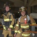 Two firefighters in full turnout gear are posed together on what appears to be a stairwell landing inside a building. The firefighter on the left wears a helmet marked 'GFI' and 'MOSS', while the one on the right wears a helmet reading 'Fort Rucker Fire-Rescue' and 'Firefighter'. Both are equipped with SCBA (Self-Contained Breathing Apparatus) gear and MSA-branded equipment.