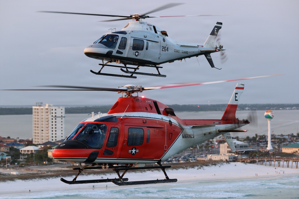 Two military training helicopters fly in formation over a coastal beach town at dusk or dawn. The upper helicopter is white and marked 'Coast Guard' with number 264, while the lower helicopter is red/orange and marked 'Marines' with number 17. Both aircraft bear the designation 'TAW' and the letter 'E' on their tail fins, indicating they are training aircraft.