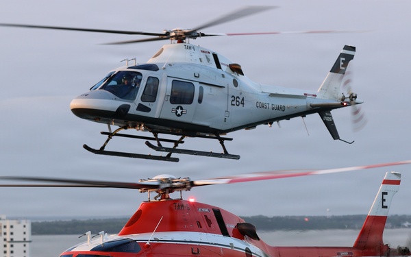 Two military training helicopters fly in formation over a coastal beach town at dusk or dawn. The upper helicopter is white and marked 'Coast Guard' with number 264, while the lower helicopter is red/orange and marked 'Marines' with number 17. Both aircraft bear the designation 'TAW' and the letter 'E' on their tail fins, indicating they are training aircraft.