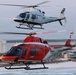 Two military training helicopters fly in formation over a coastal beach town at dusk or dawn. The upper helicopter is white and marked 'Coast Guard' with number 264, while the lower helicopter is red/orange and marked 'Marines' with number 17. Both aircraft bear the designation 'TAW' and the letter 'E' on their tail fins, indicating they are training aircraft.