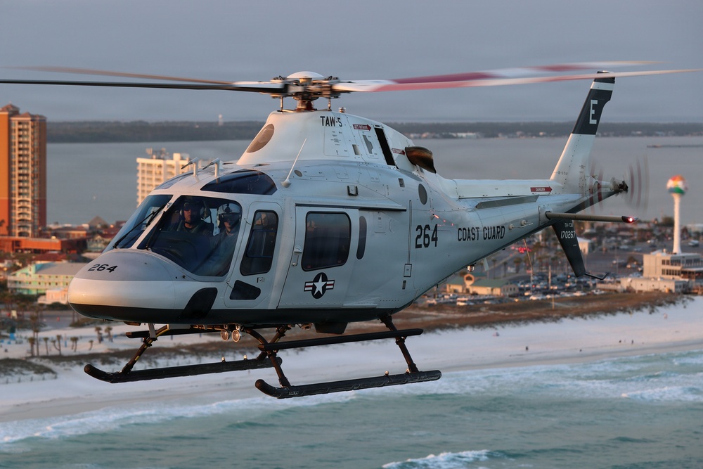 A U.S. Coast Guard helicopter numbered 264 is flying low over a coastal beach area at what appears to be dusk or dawn, given the warm golden light. The aircraft bears military markings including a star insignia and the designation 'TAW-5' on the fuselage. Two crew members are visible in the cockpit, and a coastal city with buildings and a distinctive striped tower is visible in the background.