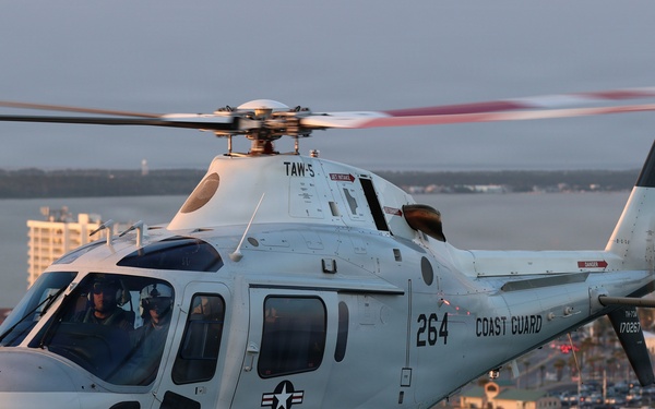 A U.S. Coast Guard helicopter numbered 264 is flying low over a coastal beach area at what appears to be dusk or dawn, given the warm golden light. The aircraft bears military markings including a star insignia and the designation 'TAW-5' on the fuselage. Two crew members are visible in the cockpit, and a coastal city with buildings and a distinctive striped tower is visible in the background.