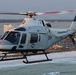 A U.S. Coast Guard helicopter numbered 264 is flying low over a coastal beach area at what appears to be dusk or dawn, given the warm golden light. The aircraft bears military markings including a star insignia and the designation 'TAW-5' on the fuselage. Two crew members are visible in the cockpit, and a coastal city with buildings and a distinctive striped tower is visible in the background.
