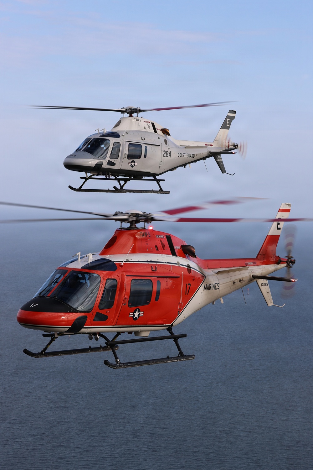 Two military training helicopters fly in formation over a body of water. The upper helicopter is white with 'COAST GUARD' and number '264' markings, while the lower helicopter is red and white with 'MARINES' and number '17' markings. Both aircraft appear to be Bell TH-57/407 or similar training variants operated by U.S. military branches.