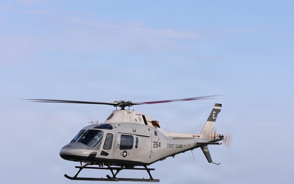 Two military training helicopters fly in formation over a body of water. The upper helicopter is white with 'COAST GUARD' and number '264' markings, while the lower helicopter is red and white with 'MARINES' and number '17' markings. Both aircraft appear to be Bell TH-57/407 or similar training variants operated by U.S. military branches.
