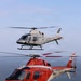 Two military training helicopters fly in formation over a body of water. The upper helicopter is white with 'COAST GUARD' and number '264' markings, while the lower helicopter is red and white with 'MARINES' and number '17' markings. Both aircraft appear to be Bell TH-57/407 or similar training variants operated by U.S. military branches.