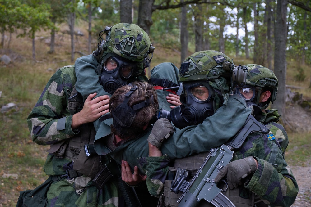 The image shows Swedish military personnel in full combat gear, including gas masks and camouflage helmets, assisting or evacuating what appears to be a casualty or incapacitated soldier. The soldiers are wearing Swedish Armed Forces uniforms identifiable by the Swedish flag patch visible on one soldier's arm. The scene appears to be a military training exercise conducted in a forested outdoor environment.