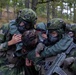 The image shows Swedish military personnel in full combat gear, including gas masks and camouflage helmets, assisting or evacuating what appears to be a casualty or incapacitated soldier. The soldiers are wearing Swedish Armed Forces uniforms identifiable by the Swedish flag patch visible on one soldier's arm. The scene appears to be a military training exercise conducted in a forested outdoor environment.