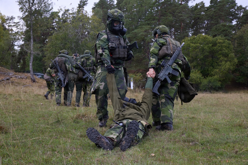 The image shows a group of soldiers in camouflage uniforms conducting what appears to be a military field training exercise in a forested outdoor area. Two soldiers in the foreground are assisting or dragging a third soldier along the ground, while additional soldiers move in the background. One soldier is wearing a gas mask and tactical vest, suggesting CBRN (Chemical, Biological, Radiological, Nuclear) training scenarios.
