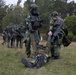 The image shows a group of soldiers in camouflage uniforms conducting what appears to be a military field training exercise in a forested outdoor area. Two soldiers in the foreground are assisting or dragging a third soldier along the ground, while additional soldiers move in the background. One soldier is wearing a gas mask and tactical vest, suggesting CBRN (Chemical, Biological, Radiological, Nuclear) training scenarios.