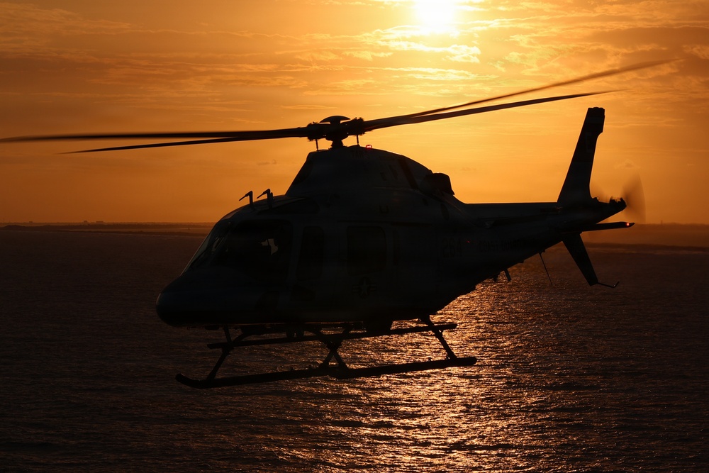A helicopter is captured in silhouette flying low over a large body of water during sunset or sunrise. The warm orange and golden tones of the sky and sun's reflection on the water create a dramatic backlit composition. The helicopter's rotor blades are visibly in motion, and the tail rotor is also spinning.