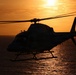 A helicopter is captured in silhouette flying low over a large body of water during sunset or sunrise. The warm orange and golden tones of the sky and sun's reflection on the water create a dramatic backlit composition. The helicopter's rotor blades are visibly in motion, and the tail rotor is also spinning.
