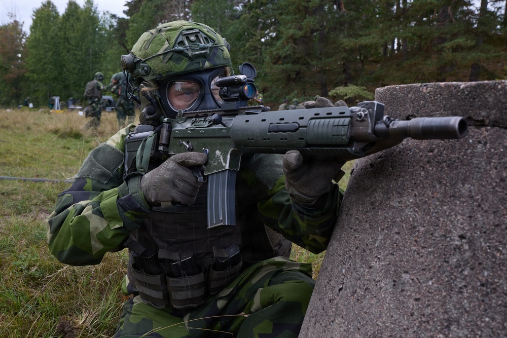 A soldier in Swedish M90 camouflage pattern uniform is taking a firing position behind a concrete barrier during what appears to be a military exercise or training scenario. The soldier is equipped with full combat gear including a gas mask, tactical helmet with rail-mounted accessories, body armor with magazine pouches, and is aiming what appears to be a Bofors/SAAB AK5 assault rifle. Additional soldiers in similar uniforms are visible in the background.