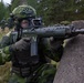 A soldier in Swedish M90 camouflage pattern uniform is taking a firing position behind a concrete barrier during what appears to be a military exercise or training scenario. The soldier is equipped with full combat gear including a gas mask, tactical helmet with rail-mounted accessories, body armor with magazine pouches, and is aiming what appears to be a Bofors/SAAB AK5 assault rifle. Additional soldiers in similar uniforms are visible in the background.