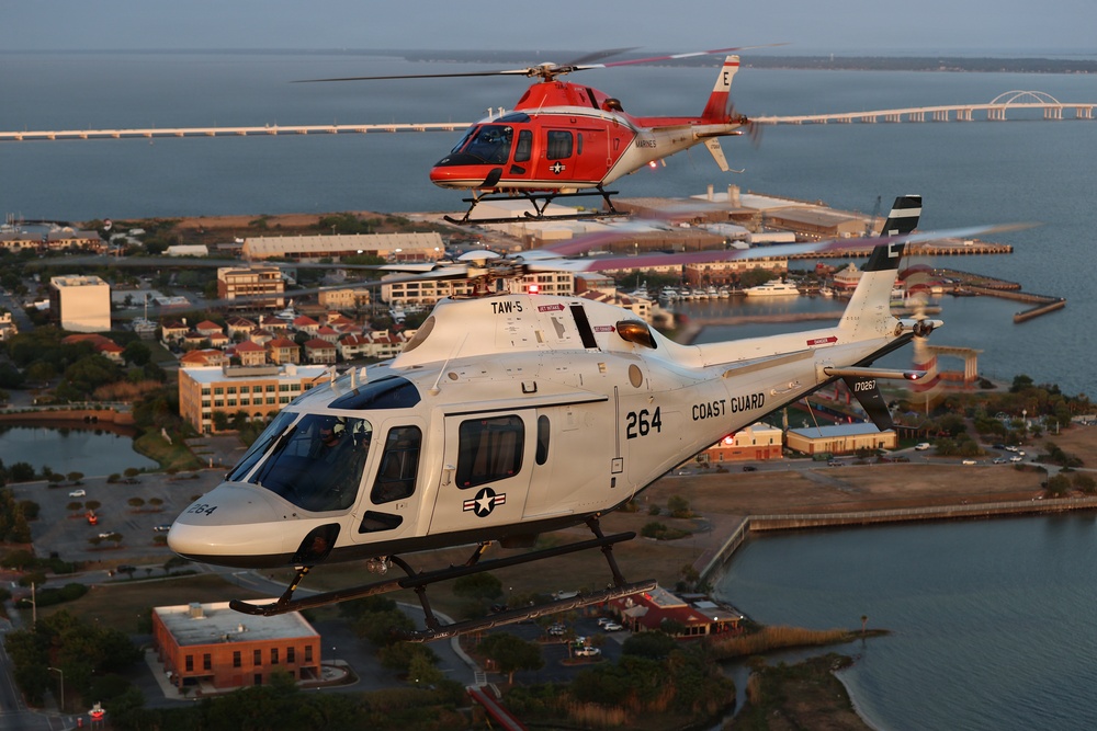 Two military helicopters are flying in formation over a coastal military installation at what appears to be dusk or dawn. The foreground helicopter is a white U.S. Coast Guard helicopter numbered 264 with markings 'TAW-5', while the background helicopter is a red and white U.S. Marines helicopter. A bridge and waterfront infrastructure are visible in the background.