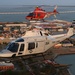 Two military helicopters are flying in formation over a coastal military installation at what appears to be dusk or dawn. The foreground helicopter is a white U.S. Coast Guard helicopter numbered 264 with markings 'TAW-5', while the background helicopter is a red and white U.S. Marines helicopter. A bridge and waterfront infrastructure are visible in the background.