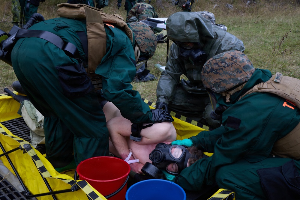 Military personnel in CBRN (Chemical, Biological, Radiological, Nuclear) protective gear are conducting a decontamination exercise on a simulated casualty. The personnel are wearing green hazmat suits, gas masks, and military helmets while treating a person lying on a yellow decontamination litter. Colored buckets (red and blue) are visible, consistent with standard decontamination station protocols.