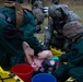 Military personnel in CBRN (Chemical, Biological, Radiological, Nuclear) protective gear are conducting a decontamination exercise on a simulated casualty. The personnel are wearing green hazmat suits, gas masks, and military helmets while treating a person lying on a yellow decontamination litter. Colored buckets (red and blue) are visible, consistent with standard decontamination station protocols.