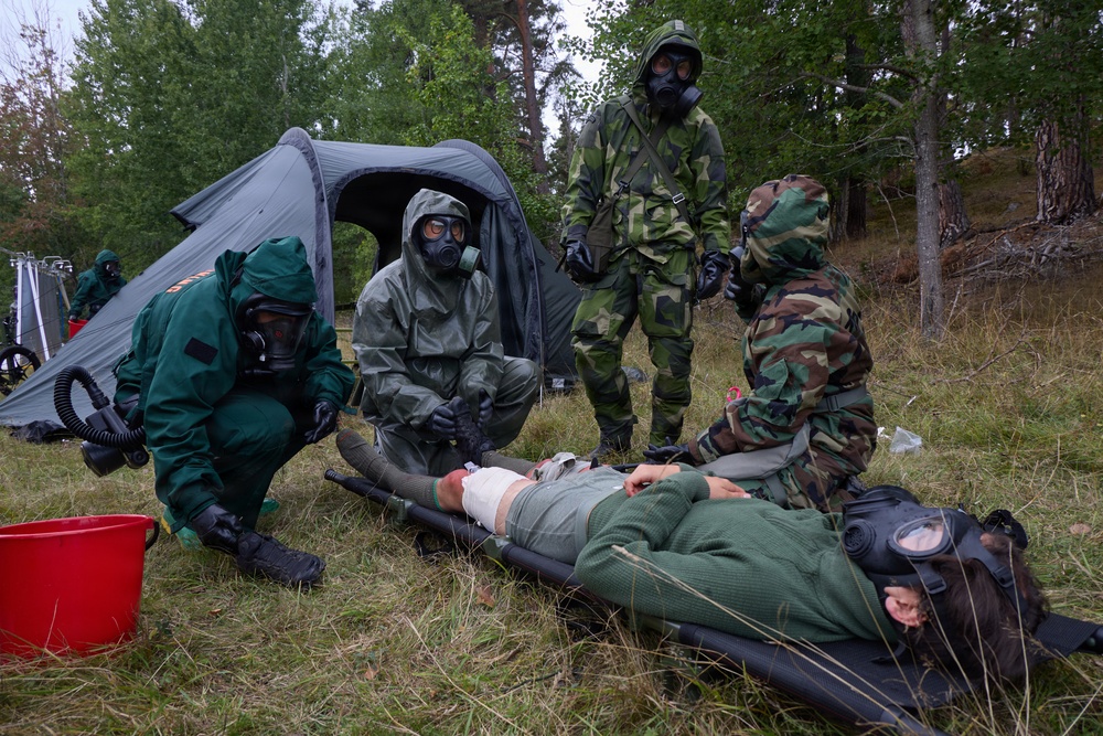A military CBRN (Chemical, Biological, Radiological, Nuclear) training exercise is taking place in a forested outdoor setting. Personnel in full protective gear including gas masks and hazmat suits are attending to a simulated casualty lying on a stretcher on the ground. The scene includes a gray military tent, medical supplies, and multiple responders in different protective suit configurations.
