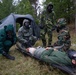 A military CBRN (Chemical, Biological, Radiological, Nuclear) training exercise is taking place in a forested outdoor setting. Personnel in full protective gear including gas masks and hazmat suits are attending to a simulated casualty lying on a stretcher on the ground. The scene includes a gray military tent, medical supplies, and multiple responders in different protective suit configurations.