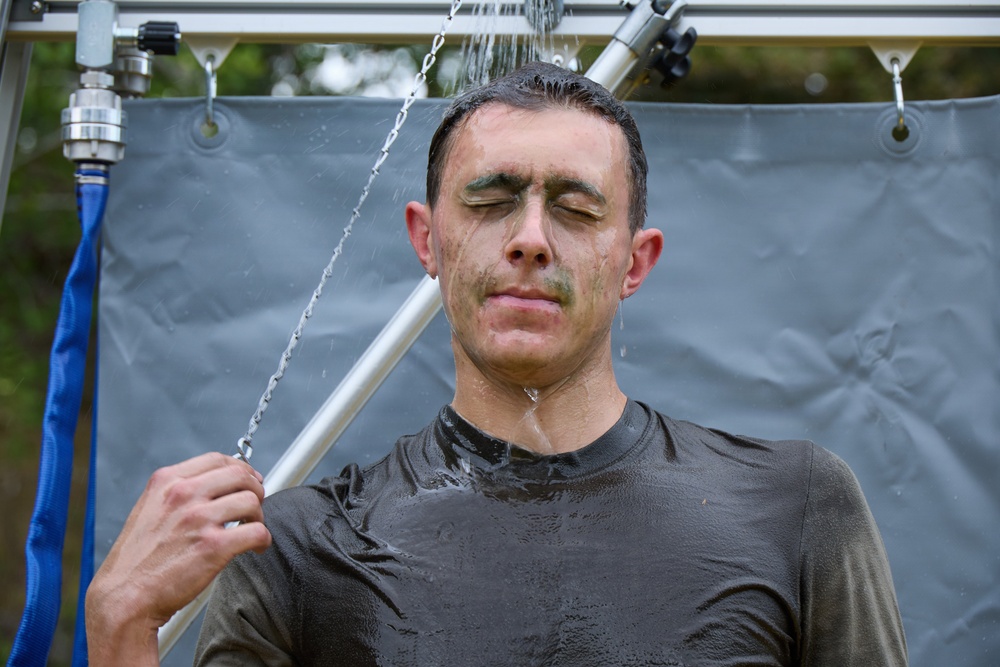 A man is standing under an outdoor shower or rinse station, with water streaming down over his head and face. He has mud or dirt on his face and clothing, suggesting he recently completed a mud run or obstacle course event. His eyes are closed and he is holding a metal bar as water washes over him.