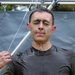 A man is standing under an outdoor shower or rinse station, with water streaming down over his head and face. He has mud or dirt on his face and clothing, suggesting he recently completed a mud run or obstacle course event. His eyes are closed and he is holding a metal bar as water washes over him.