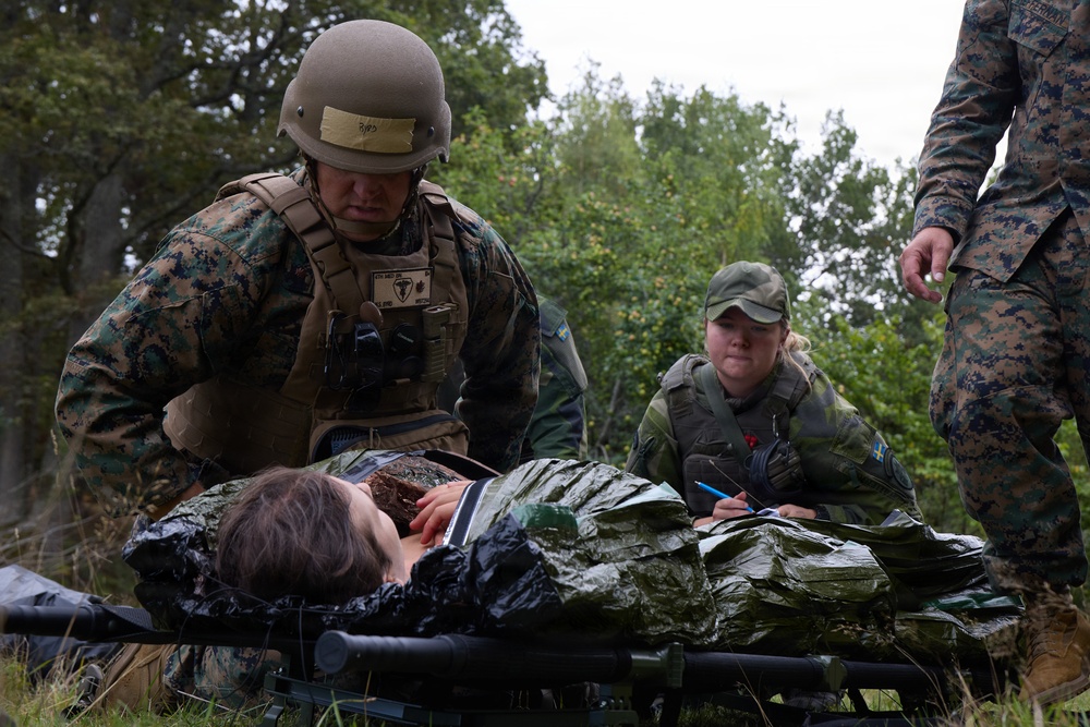 Military personnel are conducting a combat casualty care exercise, with what appears to be a simulated wounded soldier lying on a stretcher covered in a mylar/emergency blanket. A US Marine in MARPAT camouflage and combat helmet is attending to the casualty, while a Swedish military service member (identified by the Swedish flag patch) takes notes with a pen. A third military figure in MARPAT uniform is partially visible on the right side of the frame.
