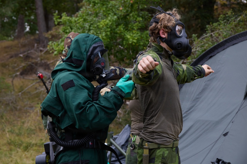 Two individuals in military or CBRN (Chemical, Biological, Radiological, Nuclear) protective gear are engaged in what appears to be a training exercise in an outdoor wooded setting. One person wearing a full green hazmat-style suit with gas mask is assisting or checking the other person, who is wearing camouflage military clothing and a gas mask. A third person in camouflage is visible in the background near a military-style tent.