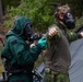Two individuals in military or CBRN (Chemical, Biological, Radiological, Nuclear) protective gear are engaged in what appears to be a training exercise in an outdoor wooded setting. One person wearing a full green hazmat-style suit with gas mask is assisting or checking the other person, who is wearing camouflage military clothing and a gas mask. A third person in camouflage is visible in the background near a military-style tent.