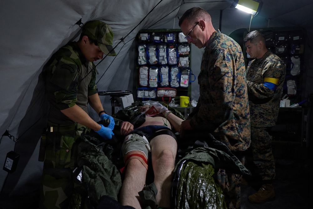 Military medical personnel from what appear to be two different armed forces are treating a casualty on a stretcher inside a field medical tent. The scene depicts a tactical combat casualty care (TCCC) scenario, likely a training exercise, with medical supplies visible on an organized wall rack in the background. Personnel wearing different camouflage patterns — one consistent with Swedish military pattern and others consistent with US Marine Corps MARPAT — are working together on the patient.