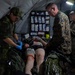 Military medical personnel from what appear to be two different armed forces are treating a casualty on a stretcher inside a field medical tent. The scene depicts a tactical combat casualty care (TCCC) scenario, likely a training exercise, with medical supplies visible on an organized wall rack in the background. Personnel wearing different camouflage patterns — one consistent with Swedish military pattern and others consistent with US Marine Corps MARPAT — are working together on the patient.