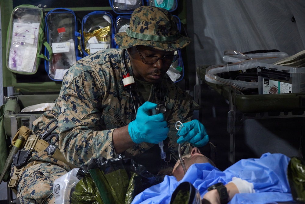 A military medic in US Marine Corps digital camouflage uniform is providing medical treatment to a patient lying on a field stretcher. The medic is wearing blue latex gloves and using a medical instrument, likely a laryngoscope or otoscope, while attending to the patient in what appears to be a forward medical treatment area or field hospital tent. Medical supplies and equipment are visible in the background.