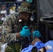 A military medic in US Marine Corps digital camouflage uniform is providing medical treatment to a patient lying on a field stretcher. The medic is wearing blue latex gloves and using a medical instrument, likely a laryngoscope or otoscope, while attending to the patient in what appears to be a forward medical treatment area or field hospital tent. Medical supplies and equipment are visible in the background.