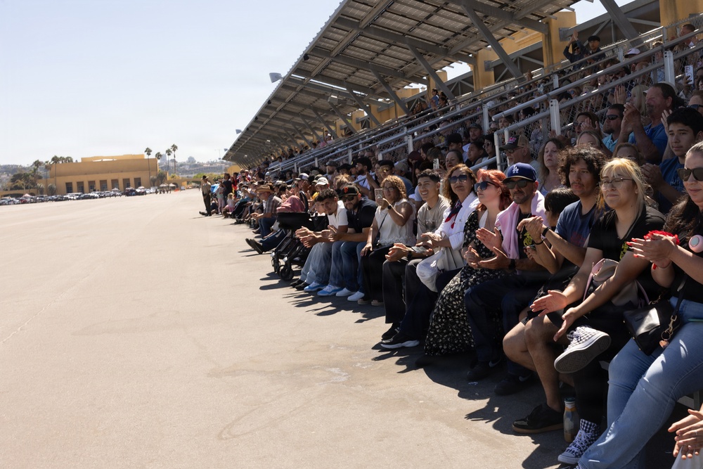 A large crowd of spectators is seated in bleachers along what appears to be a runway or tarmac at an outdoor event, likely an airshow or similar public exhibition. The audience is engaged and animated, with many people clapping, smiling, and watching something off-frame to the left. The venue features a covered grandstand with solar panels on the roof, set against a clear, sunny sky.