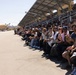A large crowd of spectators is seated in bleachers along what appears to be a runway or tarmac at an outdoor event, likely an airshow or similar public exhibition. The audience is engaged and animated, with many people clapping, smiling, and watching something off-frame to the left. The venue features a covered grandstand with solar panels on the roof, set against a clear, sunny sky.