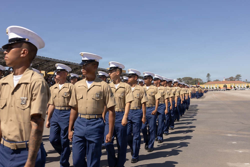A large formation of U.S. Marine Corps recruits or Marines are marching in a graduation parade on a paved drill field. They are dressed in the Service Charlie uniform consisting of khaki shirts, blue trousers, and white covers (caps). The setting appears to be a Marine Corps Recruit Depot, likely MCRD San Diego based on the visible architecture and landscape.