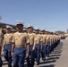 A large formation of U.S. Marine Corps recruits or Marines are marching in a graduation parade on a paved drill field. They are dressed in the Service Charlie uniform consisting of khaki shirts, blue trousers, and white covers (caps). The setting appears to be a Marine Corps Recruit Depot, likely MCRD San Diego based on the visible architecture and landscape.