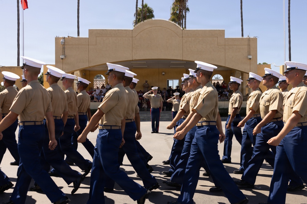 A formal military parade is taking place at what appears to be a U.S. Marine Corps base, likely MCRD (Marine Corps Recruit Depot) San Diego based on the distinctive Spanish Colonial Revival architecture. Rows of uniformed Marines in tan shirts, blue trousers, and white covers (caps) march in formation past a reviewing officer who is rendering a salute. Spectators and family members are visible in the background observing the ceremony.