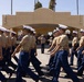 A formal military parade is taking place at what appears to be a U.S. Marine Corps base, likely MCRD (Marine Corps Recruit Depot) San Diego based on the distinctive Spanish Colonial Revival architecture. Rows of uniformed Marines in tan shirts, blue trousers, and white covers (caps) march in formation past a reviewing officer who is rendering a salute. Spectators and family members are visible in the background observing the ceremony.