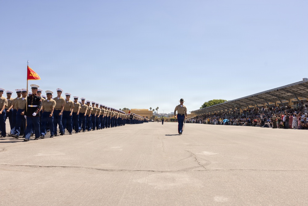 A U.S. Marine Corps graduation ceremony (boot camp graduation) is taking place on a large parade deck, likely at MCRD San Diego based on the facility layout and geography. A long formation of newly graduated Marines in tan service uniforms and white covers marches in precise columns, led by a Drill Instructor in blue trousers and campaign cover. A large crowd of spectators fills the bleachers on the right side of the parade field.