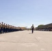 A U.S. Marine Corps graduation ceremony (boot camp graduation) is taking place on a large parade deck, likely at MCRD San Diego based on the facility layout and geography. A long formation of newly graduated Marines in tan service uniforms and white covers marches in precise columns, led by a Drill Instructor in blue trousers and campaign cover. A large crowd of spectators fills the bleachers on the right side of the parade field.