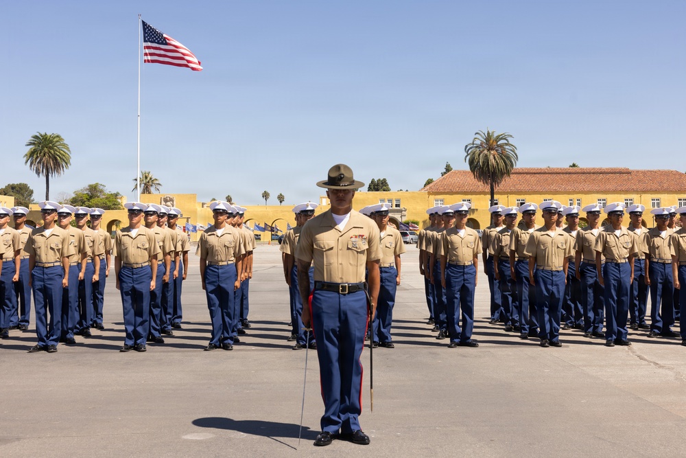 A U.S. Marine Corps drill instructor in a campaign cover (drill sergeant hat) stands at the front of a large formation of recruits or Marines on a parade deck. The personnel are dressed in the standard tan shirt and blue trousers service uniform, with white covers worn by the formation members. The setting appears to be a Marine Corps recruit depot, likely MCRD San Diego, based on the distinctive yellow Spanish Colonial Revival architecture and palm trees visible in the background.