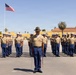 A U.S. Marine Corps drill instructor in a campaign cover (drill sergeant hat) stands at the front of a large formation of recruits or Marines on a parade deck. The personnel are dressed in the standard tan shirt and blue trousers service uniform, with white covers worn by the formation members. The setting appears to be a Marine Corps recruit depot, likely MCRD San Diego, based on the distinctive yellow Spanish Colonial Revival architecture and palm trees visible in the background.